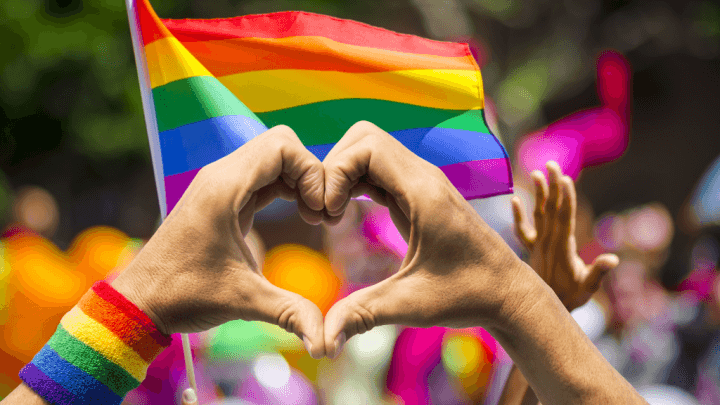manos haciendo una forma de corazón con la bandera del arco iris de fondo